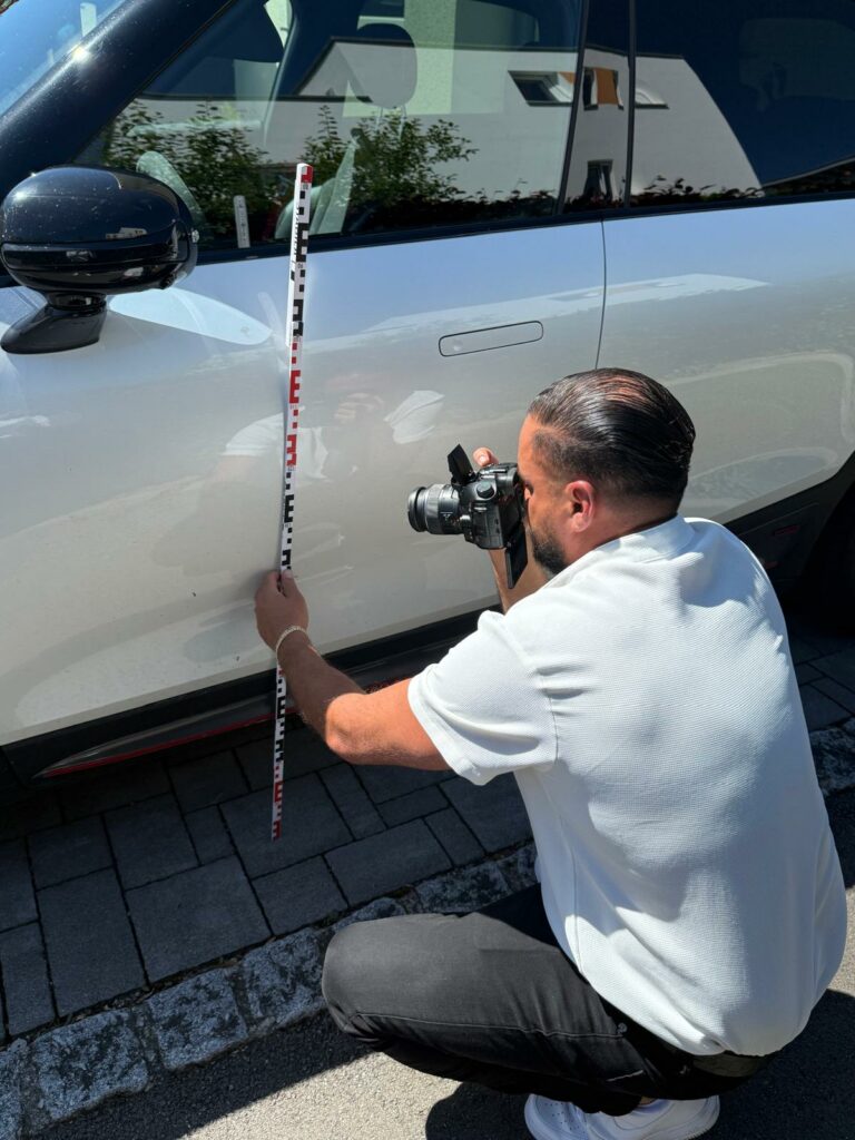 KFZ Gutachter fotografiert Seitenschaden an weißem PKW mit Kamera und Messlatte – detaillierte Schadensdokumentation für Unfallgutachten.