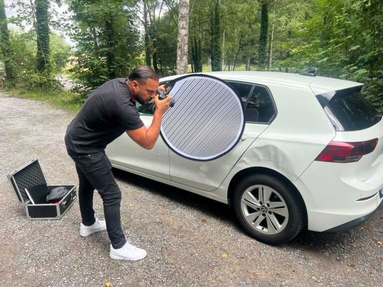KFZ Gutachter fotografiert Seitenschaden an weißem Auto mit Kamera und Reflektor – präzise Dokumentation für Unfallgutachten.