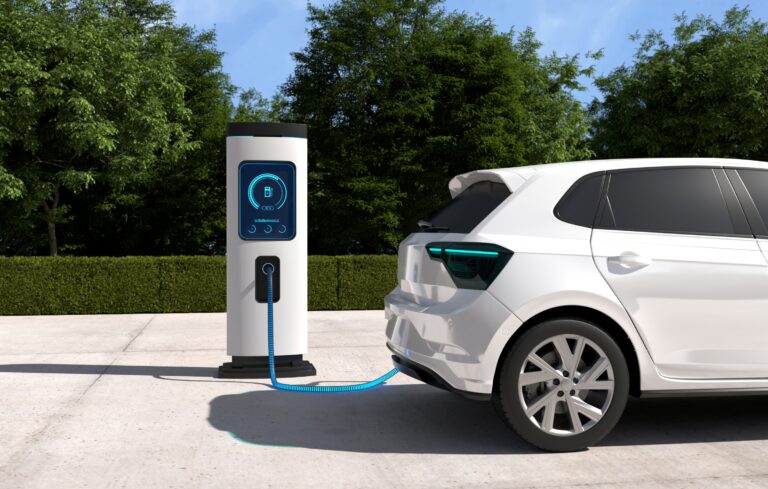 Weißes Elektroauto an moderner Ladesäule – Ladekabel eingesteckt, lädt im Freien (Parkplatz mit Bäumen im Hintergrund).