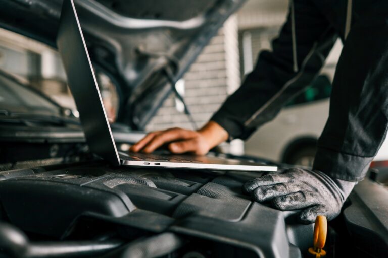 Mechaniker mit Handschuhen führt eine Fahrzeugdiagnose in Fachwerkstatt am offenen Motorraum durch – Laptop liegt auf dem Motor.