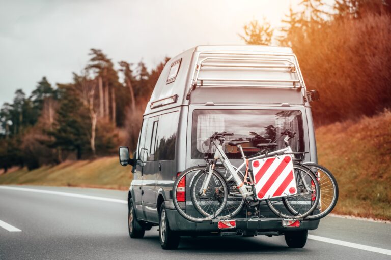Wohnmobil mit Fahrradträger (zwei Räder und rot-weißes Warntafelschild) fährt auf einer Landstraße; Bäume im Hintergrund.